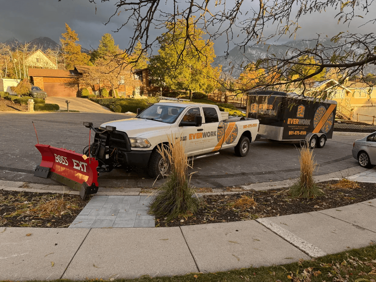 Everwork truck with a red snow plow and branded trailer parked beside landscaped beds in fall